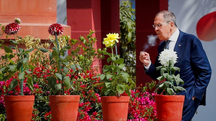 Sergei Lavrov standing behind a row of potted flowers smoking a cigarette