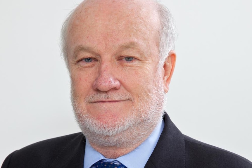 A balding white haired man is dressed in a black suit with a light blue button up and dark blue tie. He's smiling 