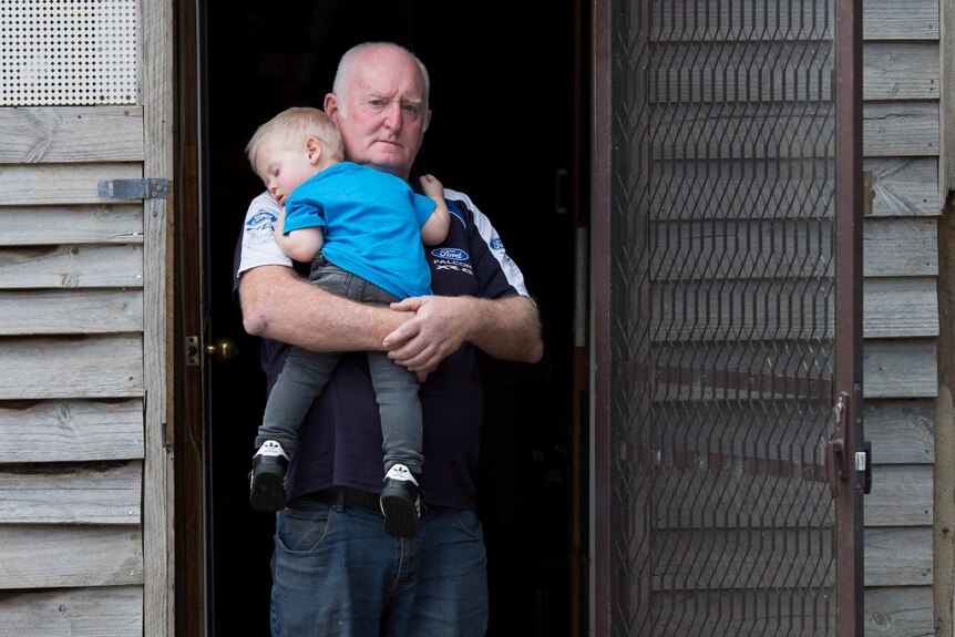 Lyndon Spicak in the doorway of his caravan annex, holding his sleeping grandson.