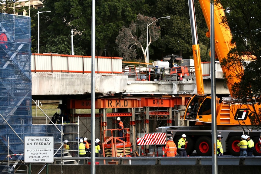 Construction workers wearing high-vis and helmets stand at the base of the Hay Street bridge with a large crane next to them.