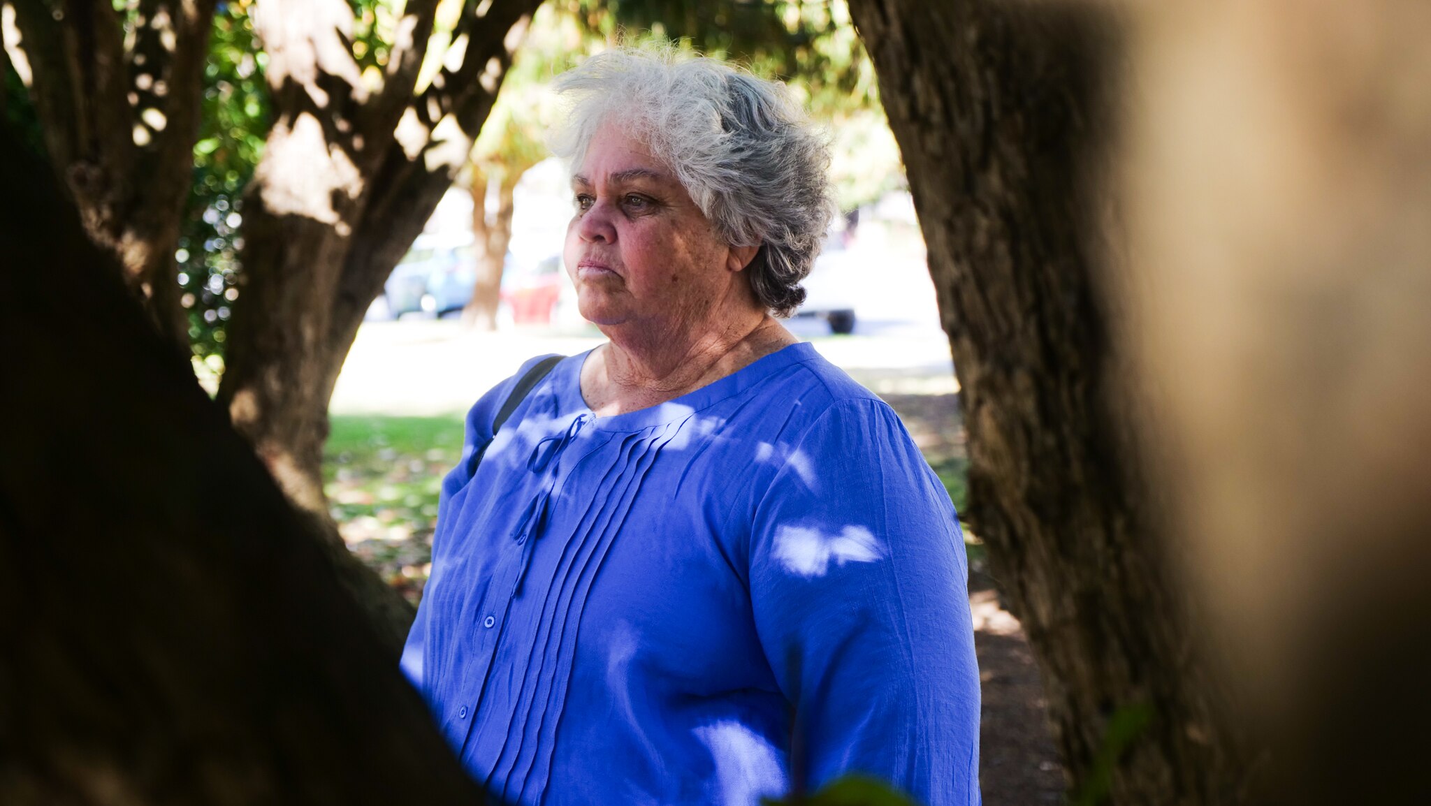 Woman in blue shirt looks out sombrely between tree branches.