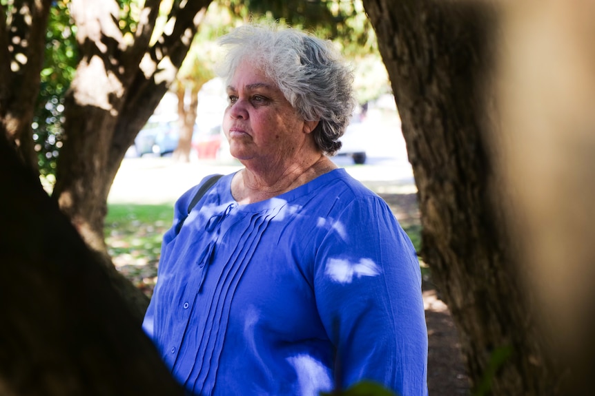 Woman in blue shirt looks out sombrely between tree branches.