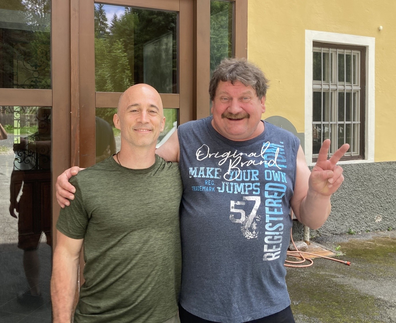 Dois homens de camiseta sorriem de braços dados em frente a uma casa.