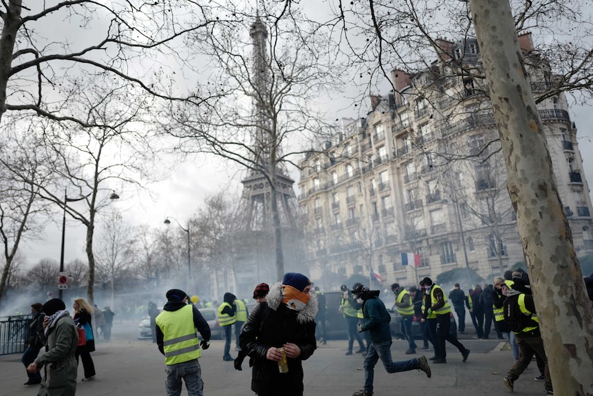 Yellow vest protesters run away from tear gas fired by riot police with the Eiffel Tower in the background.