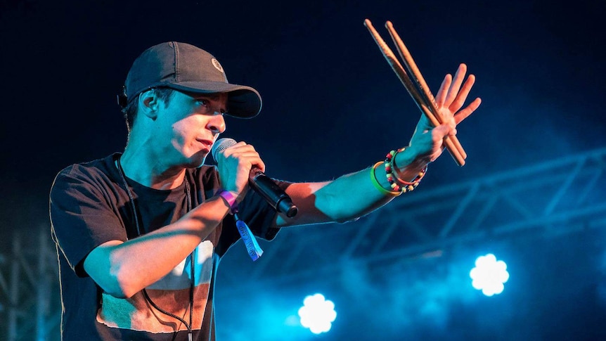 Rhyan is on stage holding a microphone to his mouth and holds two drum sticks. He wears a black cap and t-shirt.