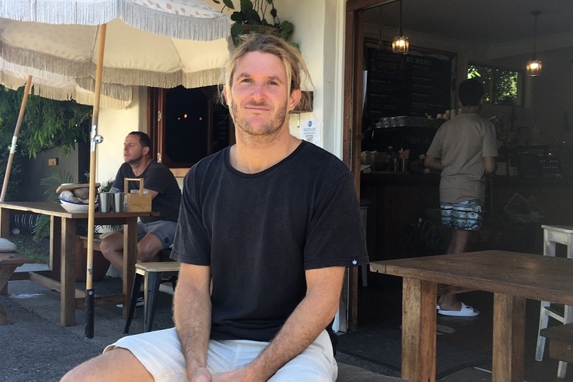 A man with long blond hair sits outside a table at a cafe.