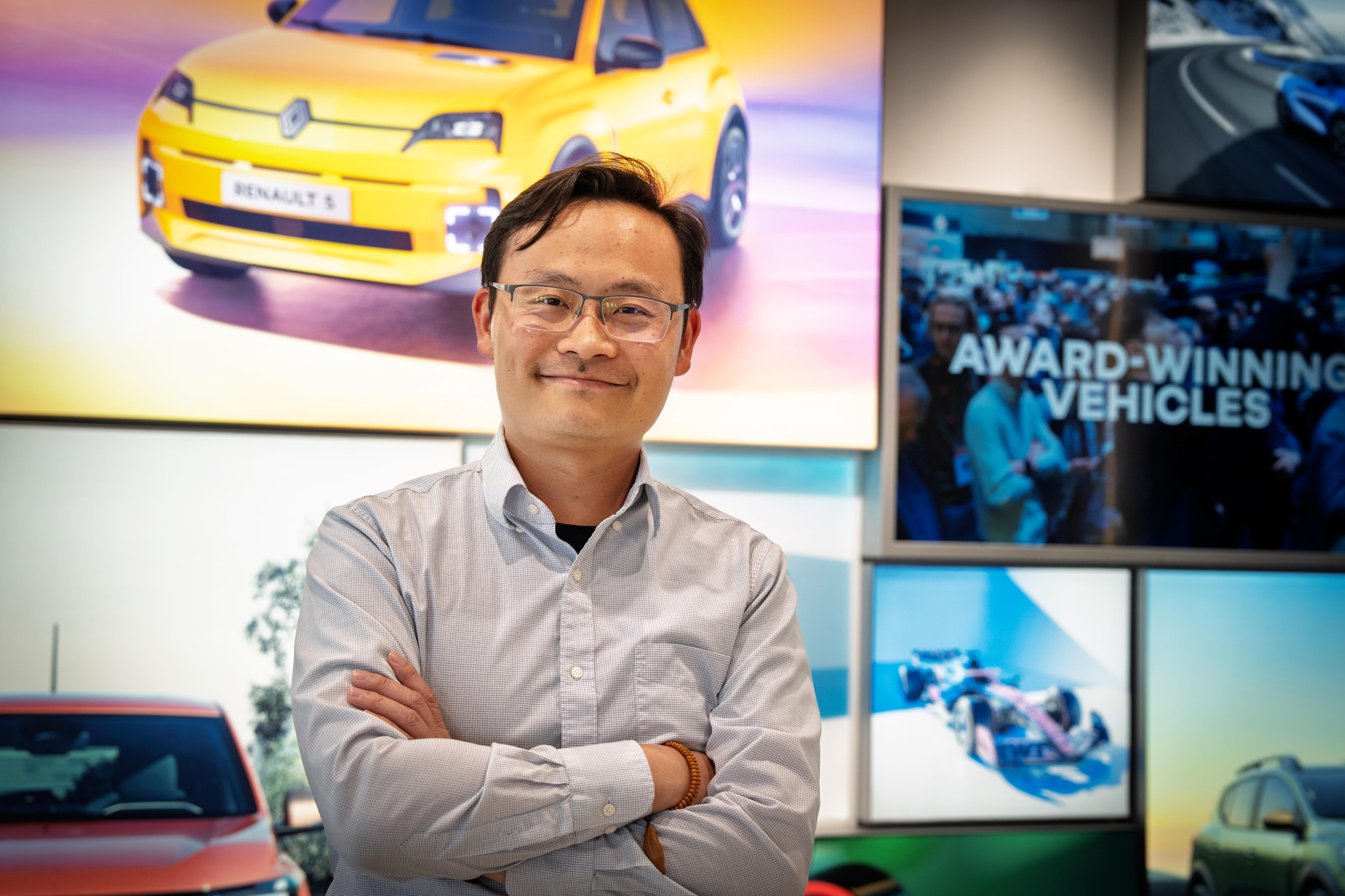 A man with glasses smiling with his arms crossed in front of screens displaying EVs.
