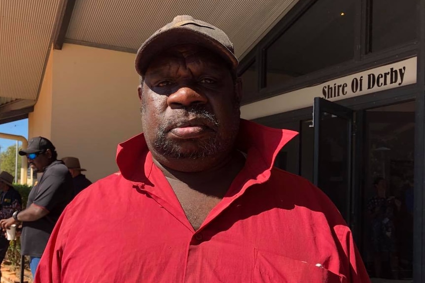 Image of a dark skinned man wearing a cap and red polo shirt, standing outside of a council building.