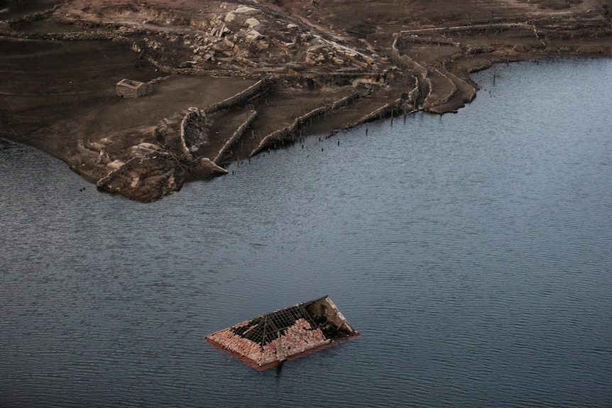 Tomados desde el aire, varios edificios revestidos de barro se asientan, en varios estados de deterioro, en un banco junto a una gran masa de agua.