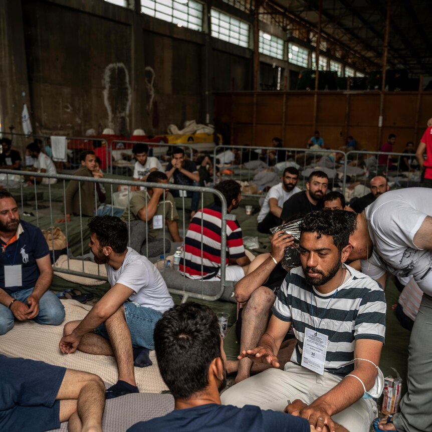Mostly male survivors of a shipwreck sit inside a warehouse, many on makeshift beds.