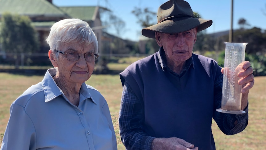 A lady in a blue shirt and a man in a blue jumper and broad brimmed hat check an empty rain gauge.