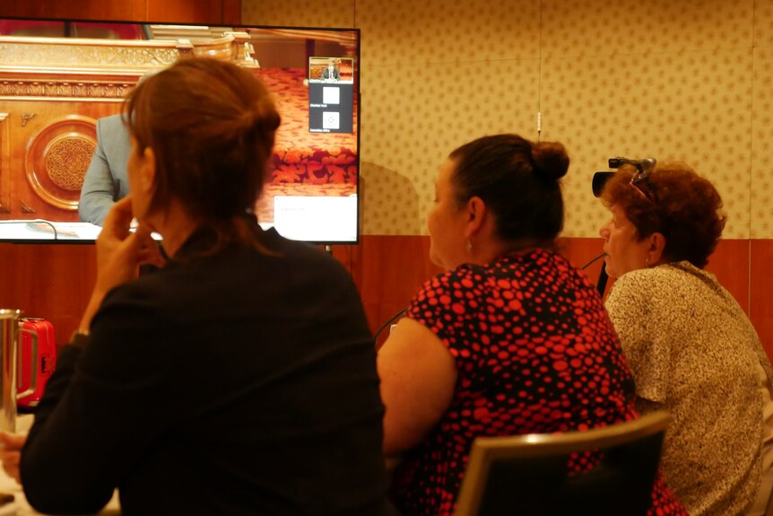 Three women sit behind a desk looking up at a screen in a boardroom.