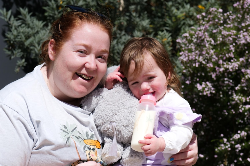 A woman smiling at the camera with her toddler daughter in her arms who is holding a bottle.