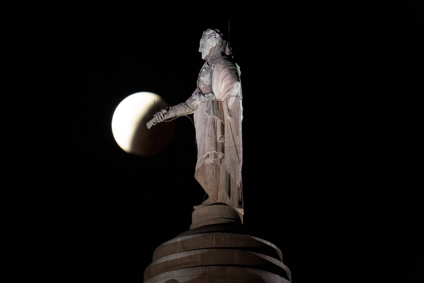 a statue of george washington is seen in the foreground at night with a lunar eclipse behind it
