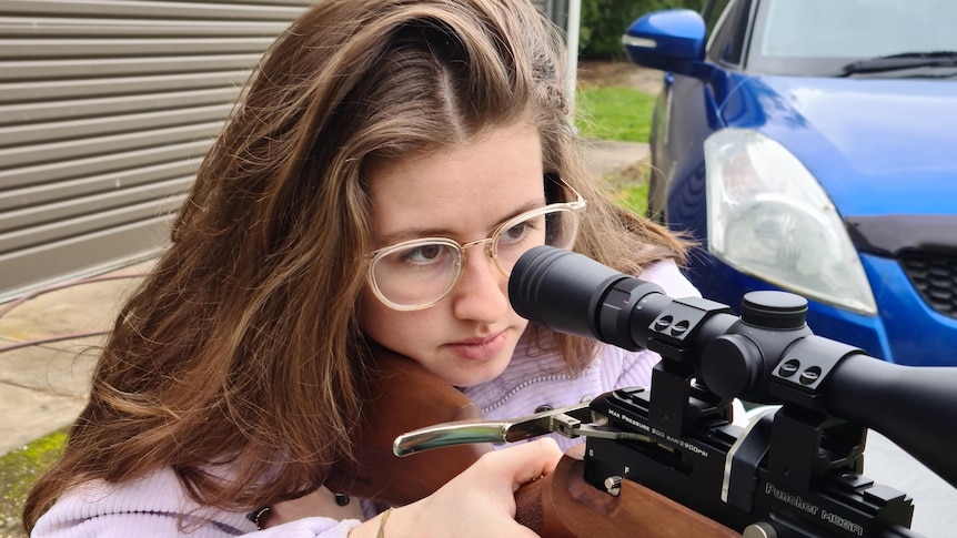 A young woman in a purple jumper aiming down the sight of a rifle