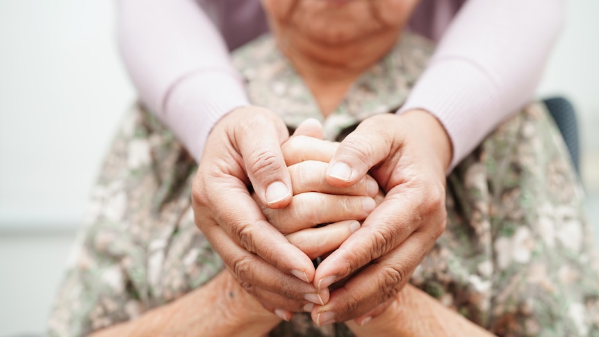 An older woman is embraced from behind by a younger woman and together their hands form the shape of a heart.