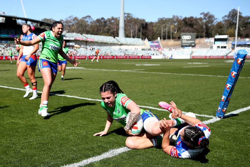 A Canberra NRLW player sits on the ground with the ball after scoring a try, as a teammate shouts in celebration.