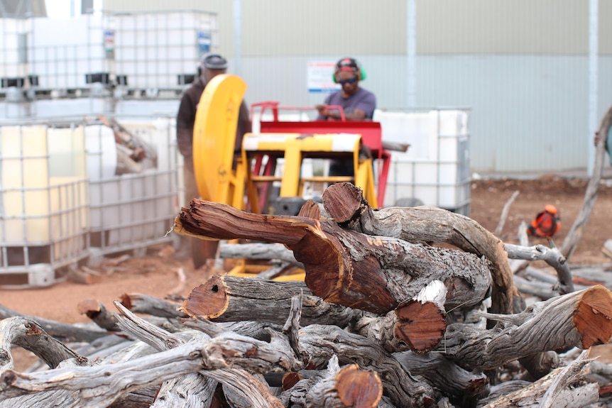 Firewood in foreground, men in background, out of focus, white bins and large yellow machinery for cutting wood
