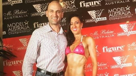 Woman in pink bikini stands with her husband in front of a competition backdrop smiling