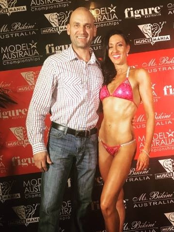 Woman in pink bikini stands with her husband in front of a competition backdrop smiling