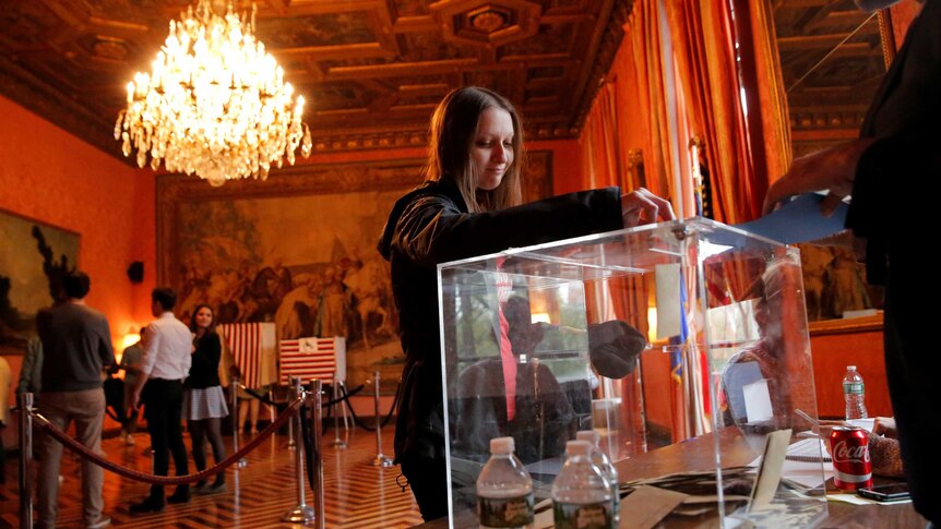 Person inserting a vote into the box, people line up behind her in an ornate setting with chandelier