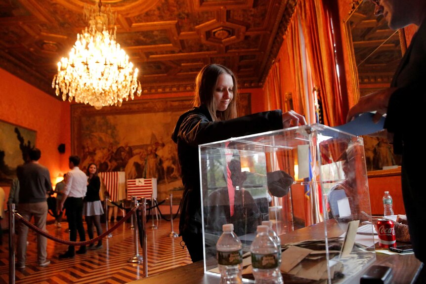 Person inserting a vote into the box, people line up behind her in an ornate setting with chandelier