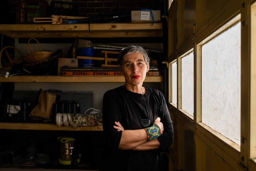 A woman wearing blacks stands with arms folded in a garage, natural lighting from window