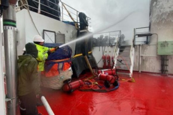 A man in a high-vis vest aims a fire hose and sprays foam onto a fire on the deck of a ship.