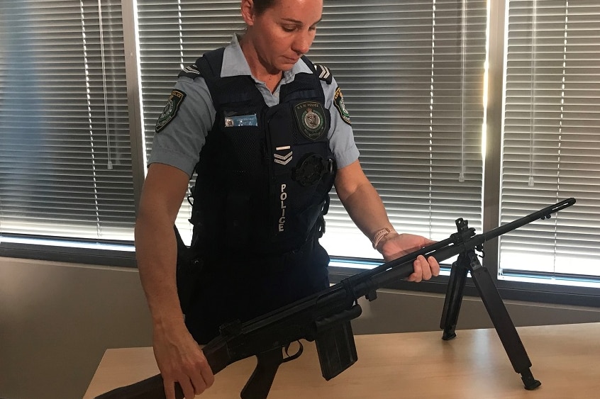 A female police officer in uniform inspects a military-grade firearm on an office desk.