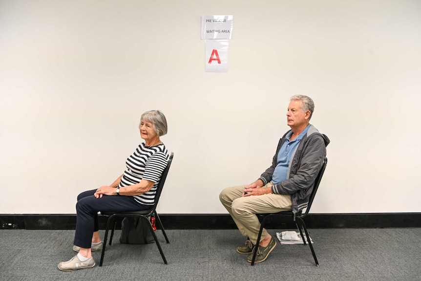 Un hombre y una mujer se sientan en dos sillas separadas por 1,5 metros en la sala de espera de vacunación.