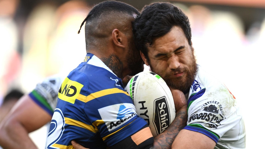 Two male NRL players are involved in a standing tackle, with one carrying the ball as he is wrapped up by the other.