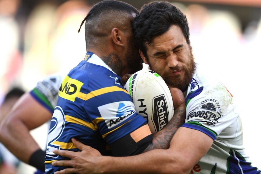 Two male NRL players are involved in a standing tackle, with one carrying the ball as he is wrapped up by the other.