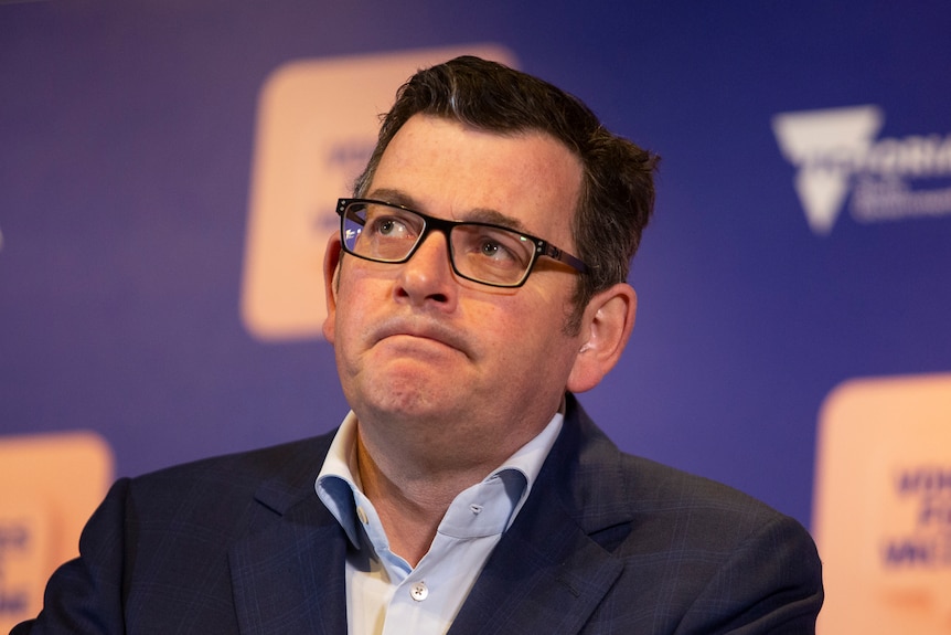 Daniel Andrews in a shirt and jacket standing at a lectern in front of a purple health department backdrop.