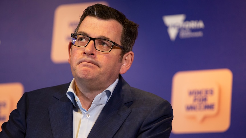 Daniel Andrews in a shirt and jacket standing at a lectern in front of a purple health department backdrop.