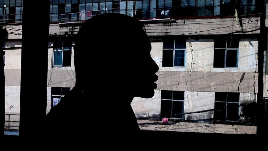 A Myanmar woman trafficked to China speaks in front of a window. She is silhouetted. There is a run-down building outside.