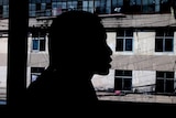 A Myanmar woman trafficked to China speaks in front of a window. She is silhouetted. There is a run-down building outside.