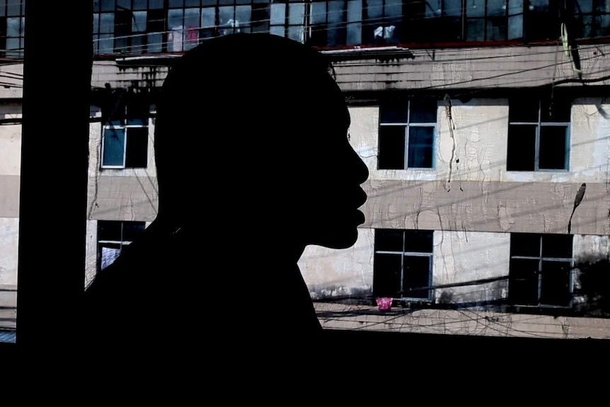 A Myanmar woman trafficked to China speaks in front of a window. She is silhouetted. There is a run-down building outside.
