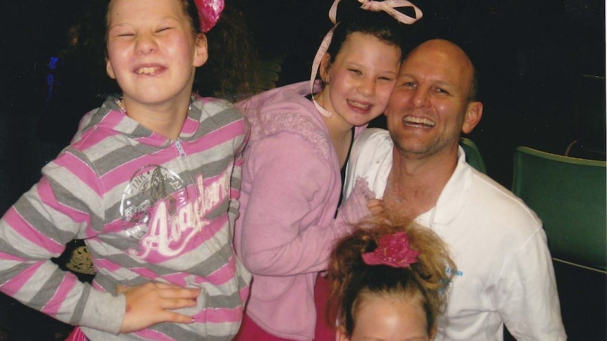 A father is surrounded by his three daughters, all wearing pink.  All have big smiles on their faces