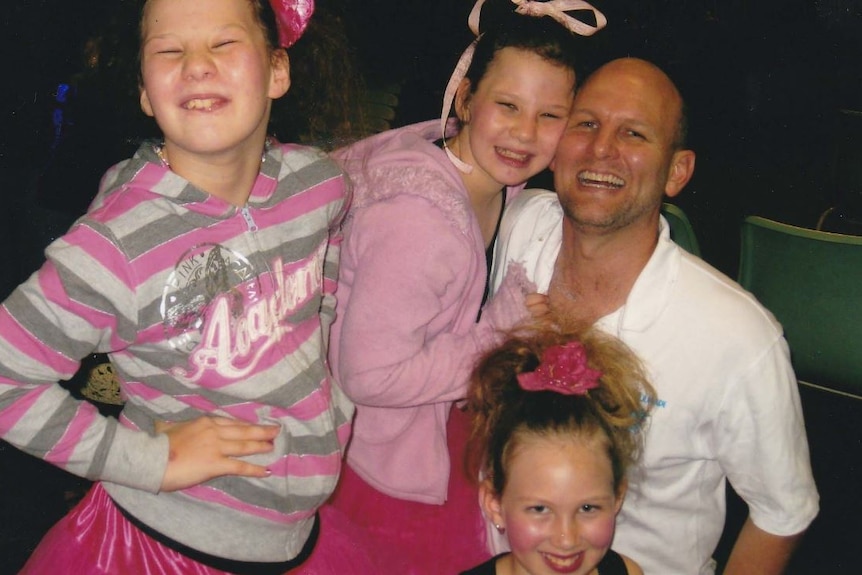 A father is surrounded by his three daughters, all wearing pink.  All have big smiles on their faces