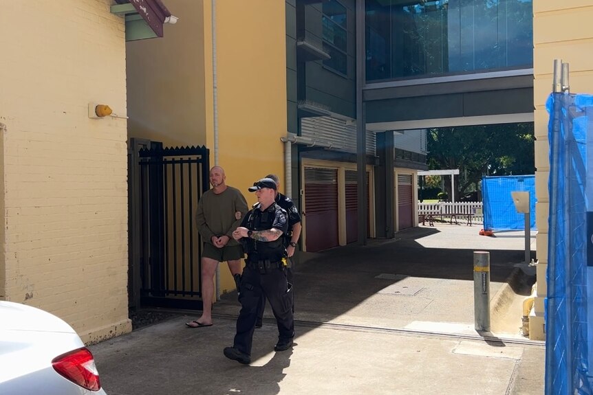 A man in a green sweatshirt wears handcuffs while being escorted by two police officers