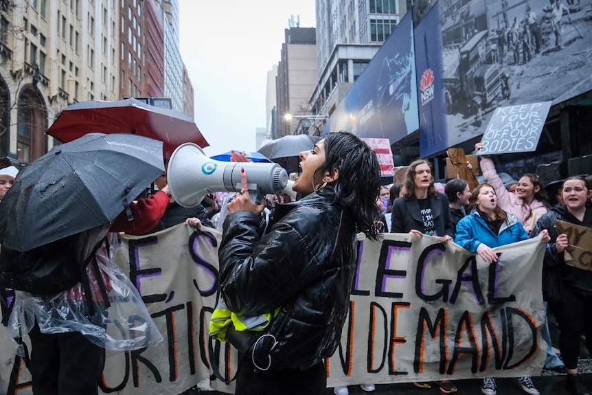 O femeie într-o jachetă neagră care vorbește prin megafon în fața protestatarilor ținând pancarte pro-choice