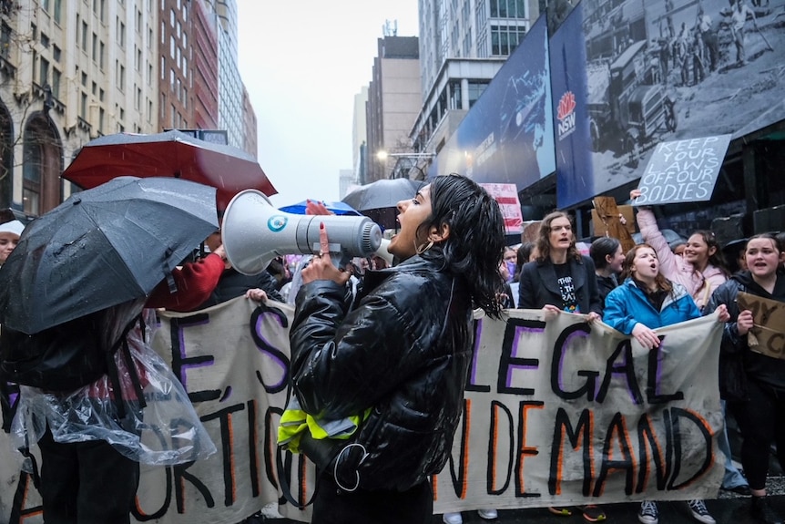 a woman in a black jacket speaks into a megaphone in front of protesters holding pro choice signs