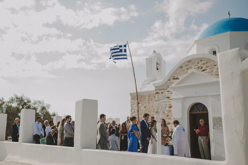 A bride and groom stand in front of a whitewashed church with a domed blue roof, there is a greek flag, a priest and a crowd