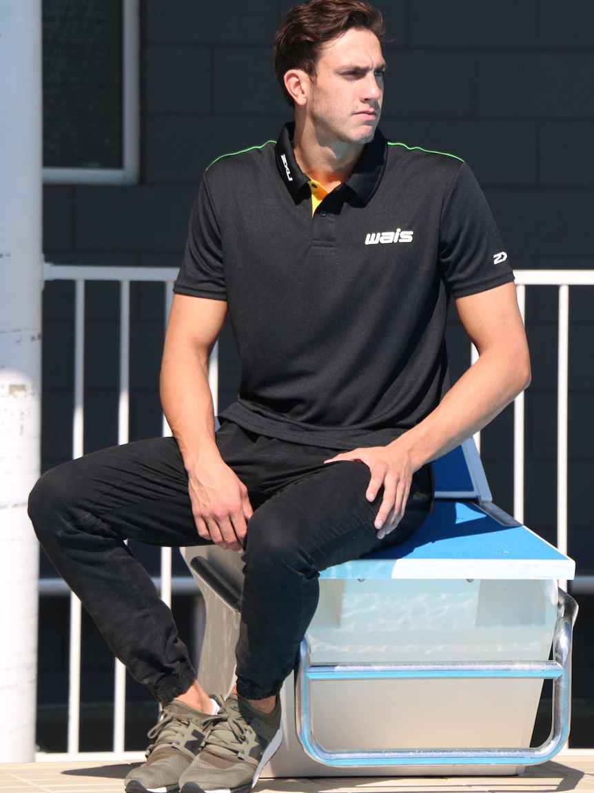 A man in black trousers and a black WAIS shirt sits on swimming blocks at an outdoor pool.