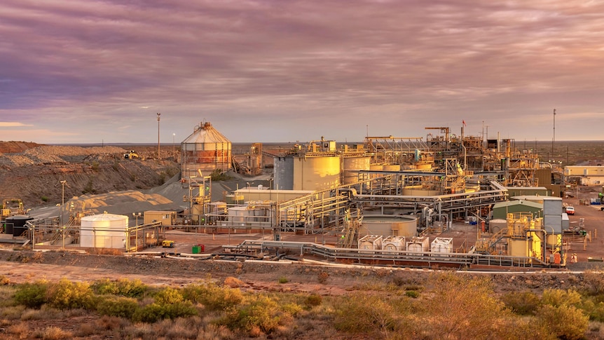 A wide shot shows facilities at a gold mine, as the sun sets and turns the sky purple