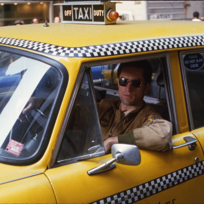 A man in sunglasses and brown jacket sits behind the wheel of a yellow taxi.