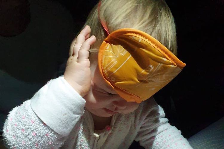 A baby wearing an eye mask on her head and looking unhappy.