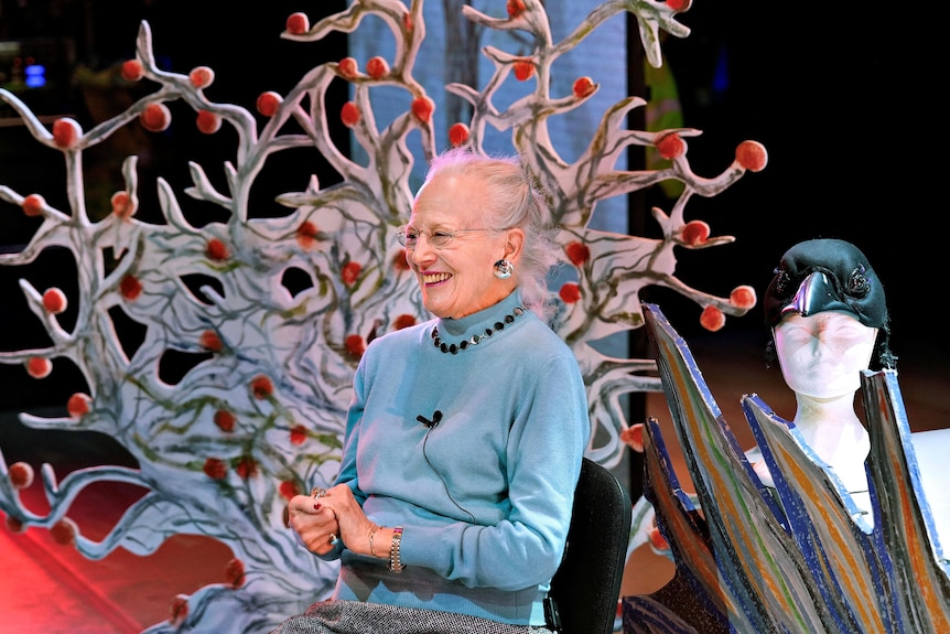 An older woman in blue jumper smiles on stage with sets from a ballet