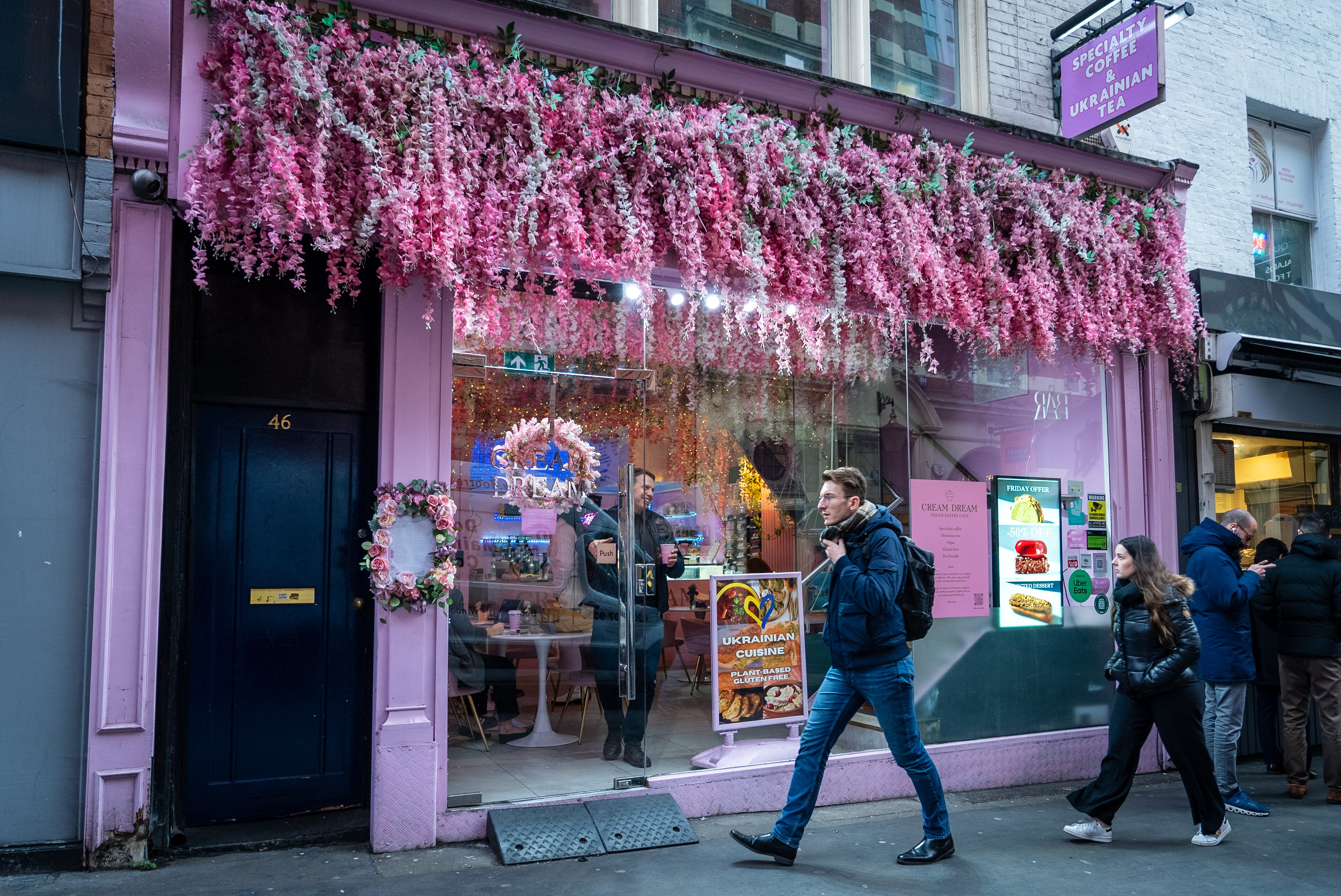People walk past a pink cafe with pink flowers hanging from the front window