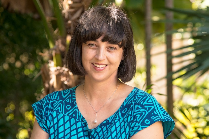 A woman smiles at the camera, large tropical plants in background.